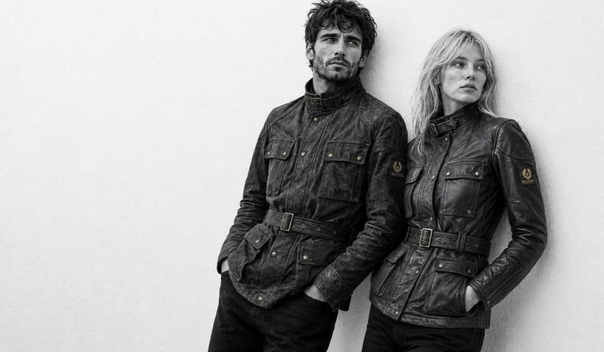 Black-and-white editorial portrait of a male and female model leaning against a light wall, both wearing worn belted field jackets in a Belstaff Trialmaster style.