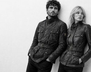 Black-and-white editorial portrait of a male and female model leaning against a light wall, both wearing worn belted field jackets in a Belstaff Trialmaster style.