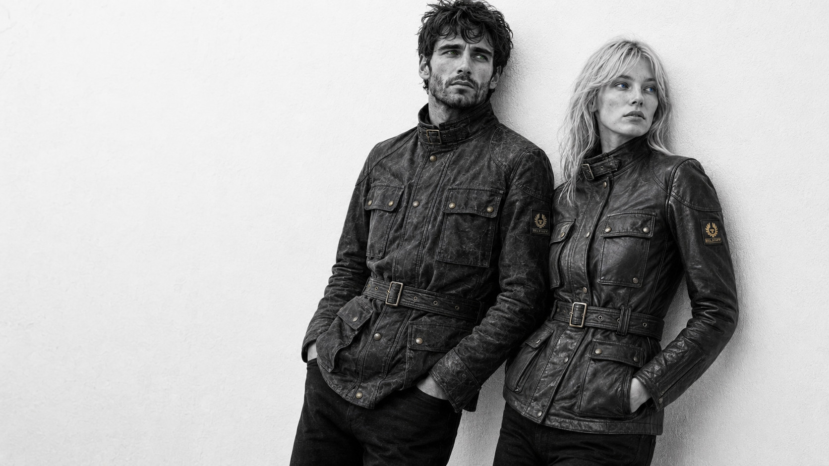 Black-and-white editorial portrait of a male and female model leaning against a light wall, both wearing worn belted field jackets in a Belstaff Trialmaster style.