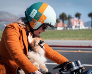 Woman riding a motorcycle during The Distinguished Gentleman’s Ride with a small dog secured in a harness, both wearing helmets, photographed on a sunny coastal road.