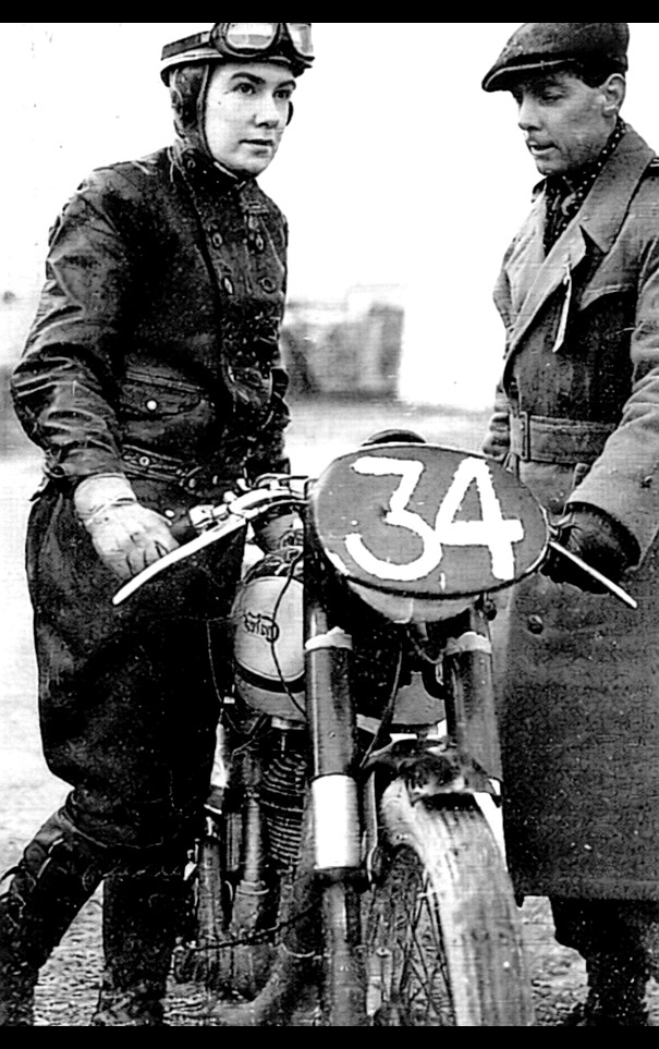 Black-and-white photograph of Olga Kevelos wearing leather racing gear, standing outdoors next to her Norton International motorcycle before a race.