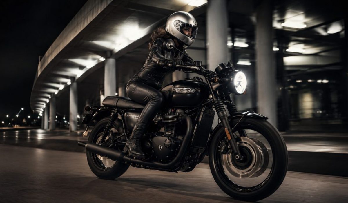 Female rider in a one-piece leather suit beside a black Triumph Bonneville T120-style motorcycle under urban night lights.