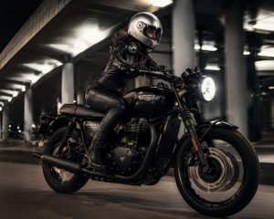 Female rider in a one-piece leather suit beside a black Triumph Bonneville T120-style motorcycle under urban night lights.